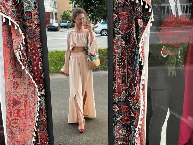 Alyce Erway, con i capelli grigi ricci, indossa una canotta rosa e pantaloni a vita alta dello stesso colore, ed è in piedi di fronte a uno specchio con tende a motivi rossi e neri..