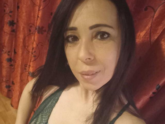 LauraSin, com cabelo roxo escuro, olhos castanhos e um sorriso discreto, vestindo uma blusa verde de renda, mostrando seus pequenos seios, em frente a uma cortina vermelha com padrões florais pretos..