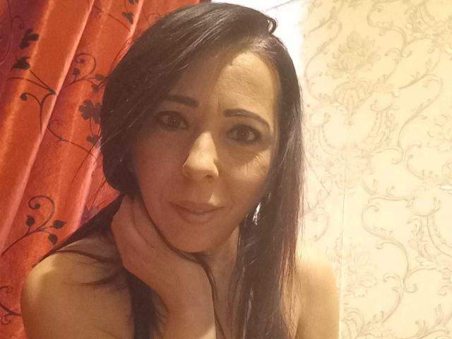 LauraSin tem cabelo castanho escuro, pele de tom médio, está tocando o pescoço com uma mão, usando um colar, com um leve sorriso e olhando para a câmera..