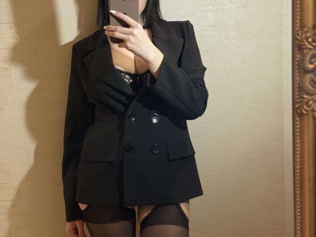 EmmaNoa in a black blazer, exposing her tits in a lace bra, wearing black garter stockings, holding a phone, against a beige wall.