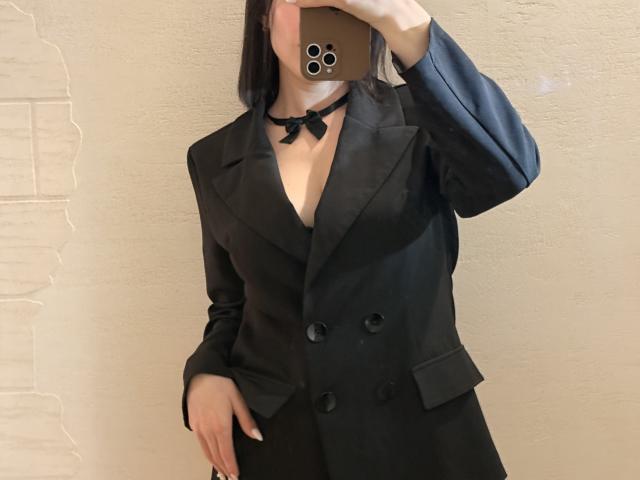 EmmaNoa with black blazer, showing cleavage, black choker, and holding phone covering face. She's standing, hand on hip, phone has three camera lenses. Noa's tits are visible.