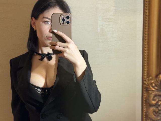 EmmaNoa with black hair, wearing a black blazer and lace bra, showing her tits in a mirror selfie, holding a gold iPhone, choker with a bow.