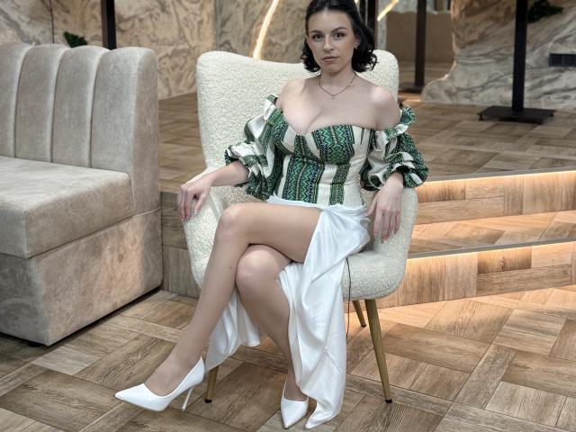 EmmaNoa with her tits showing in a green and white off-shoulder dress, white high heels, and a white skirt, sitting on a chair, legs crossed.