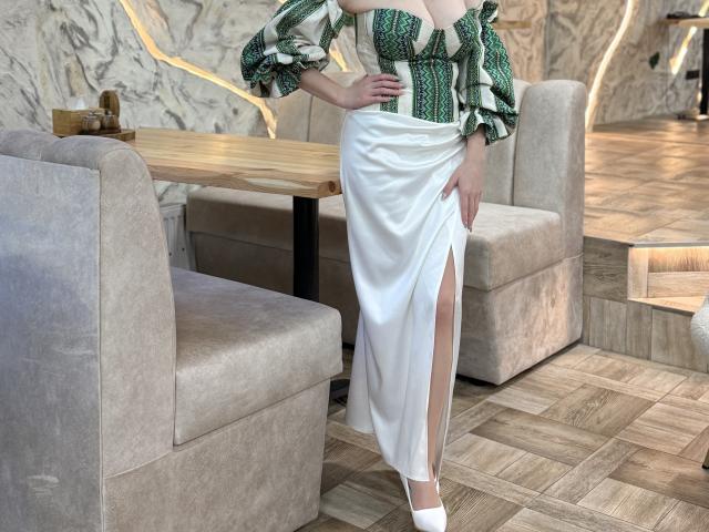 EmmaNoa in a green and white patterned top with puffed sleeves, wearing a white satin skirt with a thigh-high slit, standing in a room with beige chairs.