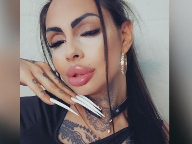 DenyChantal has dark hair, thick eyebrows, pink glossy lips, tattoos, long white nails, black choker, and gold earrings, with a sexy, confident expression.