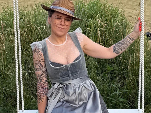 MiaHase, aantrekkelijk met een grijze jurk, zichtbare borsten, tatoeages op de arm, een bril, een strohoed, zittend op een witte schommel, een touw vasthoudend, een grasachtige achtergrond, rode nagels, lachend..