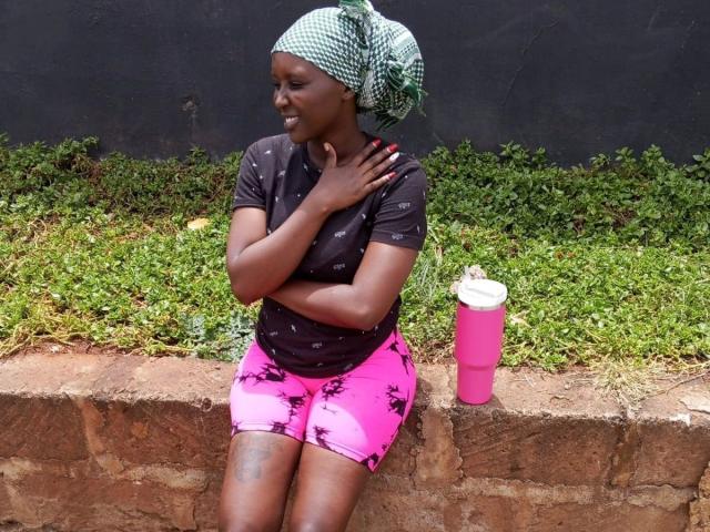 EbonyBella-hot with dark skin, smiling, wearing a green checkered headscarf, black top, pink shorts with black patterns, sitting on a brick wall, pink water bottle to her right.