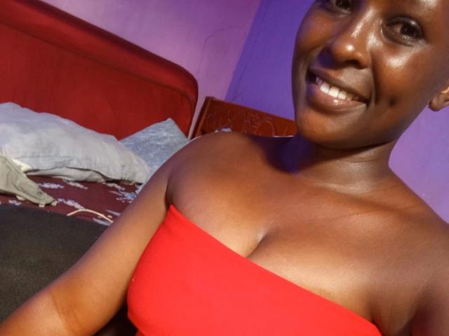 EbonyBella-hot with dark skin, smiling, wearing a red strapless top, showing her tits, lying on a bed with pillows. She's looking at the camera, showing white teeth.