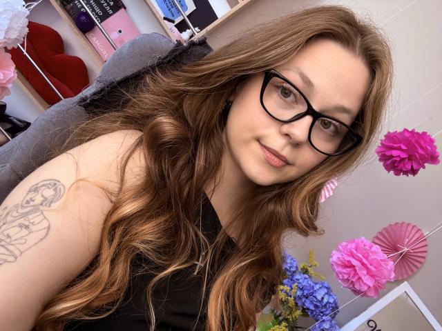 KarKutie met lang, bruin golvend haar, zwarte bril, zwarte top, tatoeage op de rechterarm, lichte glimlach, zittend, roze en roze papieren bloemen op de achtergrond..