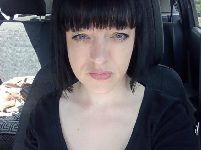 Liliit has pale skin, black bob with straight bangs, blue eyes, and pink lips. She's wearing a black V-neck shirt, sitting in a car with a blanket.