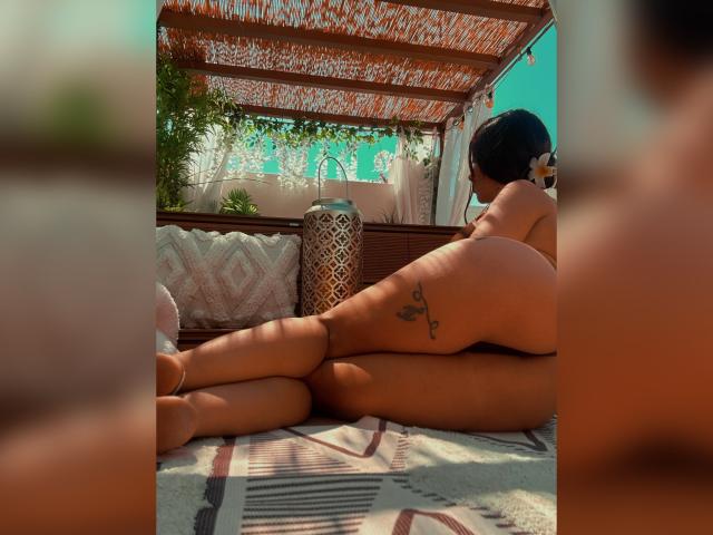 LaJoker, nue, allongée sur un tapis à motifs, les jambes croisées, tatouage sur la cuisse droite, dos à la caméra, éclairée par le soleil, au-dessus, une structure en bois, premier plan flou..