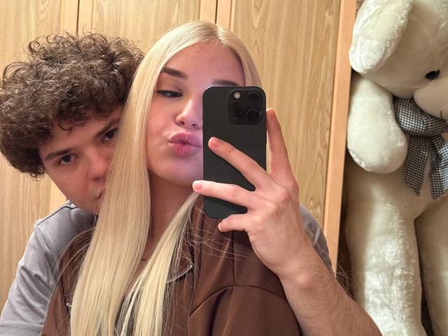 Blonde LessieKirtner avec des lèvres pulpeuses, un homme brun aux cheveux bouclés l'embrassant dans le cou, tenant un téléphone noir dans une photo prise dans un miroir, fond en bois, ours en peluche blanc avec un nœud à carreaux sur la droite..