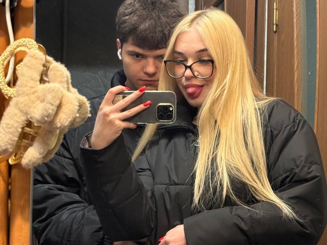 Blonde LessieKirtner with long hair and glasses, sticking tongue out, takes selfie with black puffy jacket, red nails, standing next to brown-haired guy, both in mirror.