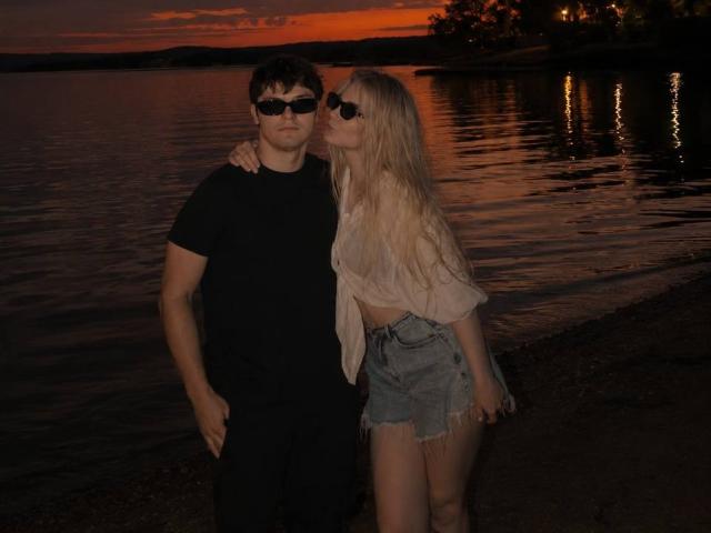 Una foto di Lessie Kirtner con i capelli biondi, che indossa occhiali da sole, una camicia annodata e pantaloncini di jeans, in piedi sulla riva, al tramonto, con una mano sulla spalla di un ragazzo..