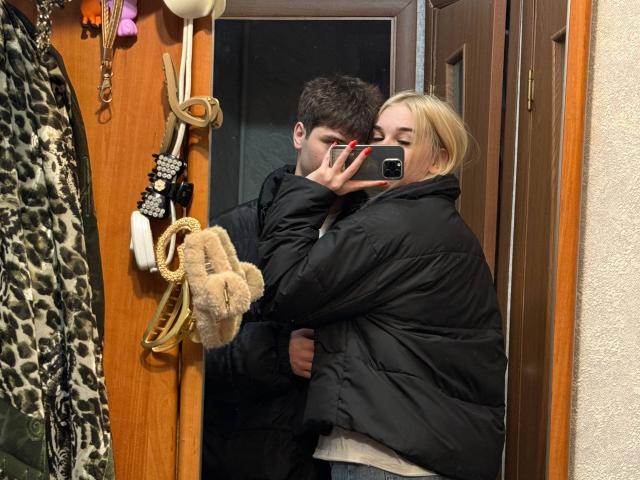 Spiegelfoto van LessieKirtner en een man in een houten gang. LessieKirtner heeft blond haar, een zwarte, dikke jas aan, houdt een telefoon vast en laat haar rug zien. De man heeft donker haar en een zwarte jas aan..