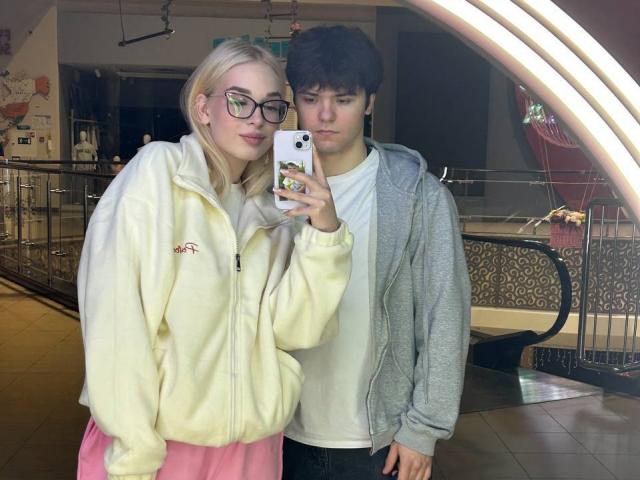 A selfie of two people, a blonde woman in a yellow fleece and pink skirt, holding a phone, standing next to a dark-haired man in a gray hoodie.