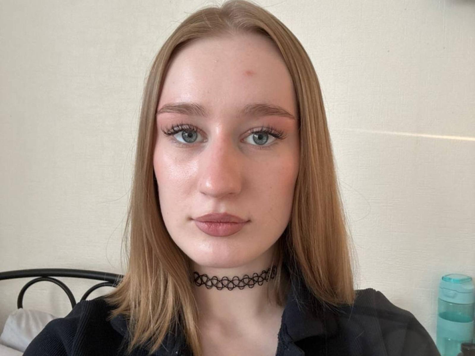 YettaMoulinos hat eine helle Haut, blaue Augen und hellbraunes Haar. Sie trägt eine schwarze Halskette und ein dunkles Hemd. Sie hat einen kleinen roten Punkt auf ihrer Stirn..
