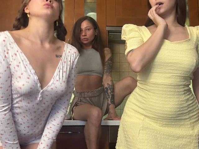Three HotCaramelsTrio standing in a kitchen. Left: white dress, small tits, pouty lips, tattoo on chest. Center: grey crop top, bare ass, tattooed arm, sitting. Right: yellow dress, pensive.
