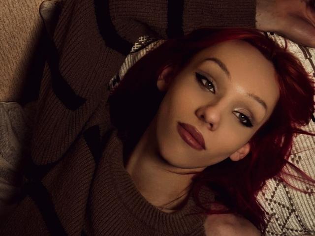 KiranaHayes, con i capelli rossi, gli occhi marroni e il rossetto scuro, sdraiata, indossa un maglione a righe marrone e nero, e guarda la fotocamera con un leggero sorriso..