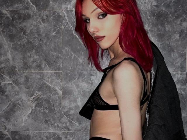 KiranaHayes, con cabello rojo brillante, un sujetador de malla negra y una chaqueta negra, mirando por encima del hombro con una sonrisa seductora, mostrando sus pequeños senos y su cuerpo delgado y pálido..