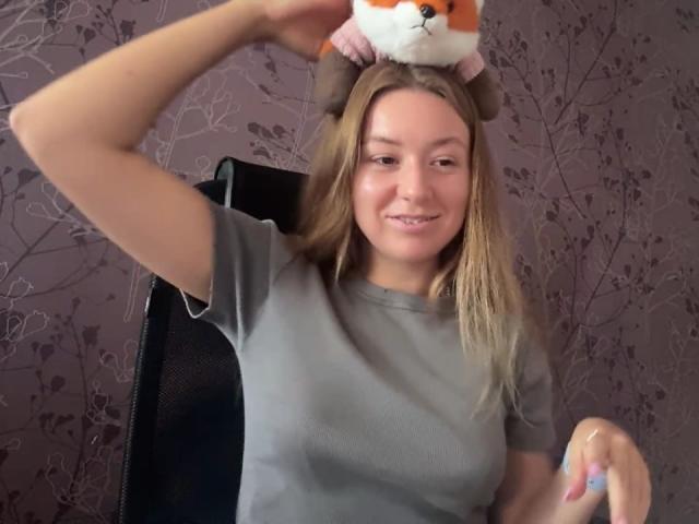 Ardelia Amesbury, con i capelli biondi, una maglietta grigia, che sorride, tiene in testa un peluche a forma di volpe, seduta su una sedia nera, braccio destro alzato, mano sinistra con le unghie rosa..