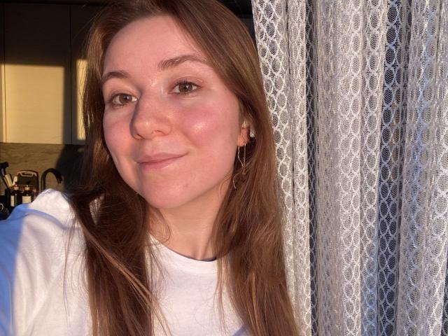 Ardelia Amesbury, con i capelli castano chiaro, un sorriso delicato, pelle rosata, indossa una camicia bianca, è in piedi alla luce del sole accanto a tende di pizzo, ha piccoli orecchini a cerchio, luce naturale soffusa..
