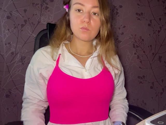 Ardelia Amesbury, com seios médios, vestindo uma regata rosa vibrante, uma camisa branca, um colar de pérolas e um prendedor de cabelo rosa, sentada em uma cadeira, olhando diretamente para frente, com uma expressão emburrada..