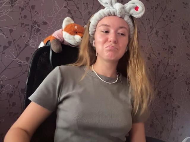 Ardelia Amesbury mit blonden Haaren, grauem Stirnband, kleinen Brüsten, grauem T-Shirt, einer Halskette, geschürzten Lippen, einem Stofffuchs auf einem Stuhl, vor einer violetten Blumenwand..