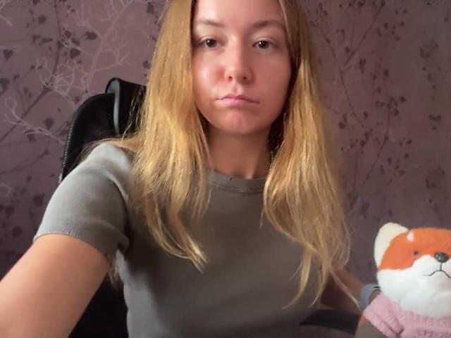 Ardelia Amesbury mit langen blonden Haaren, die ein graues Hemd trägt, mit leicht geschürzten Lippen, sitzend auf einem Stuhl, wobei der rechte Arm eine rote und weiße Stoffpuppe in Form eines Fuchses hält..