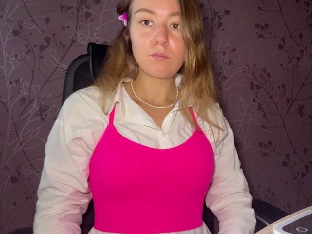 Ardelia Amesbury, con capelli biondi, canottiera rosa, camicia bianca, collana di perle e molletta rosa per capelli, seduta su una sedia, con un'espressione seria, con il seno visibile a causa della canottiera aderente..