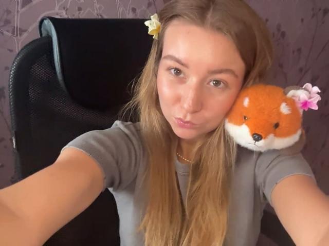 Ardelia Amesbury mit langen braunen Haaren, die ein graues Hemd trägt, verzog den Mund und posiert mit einem flauschigen, roten Fuchsplüschtier mit rosa Blumen auf ihrer Schulter in einem Selfie..