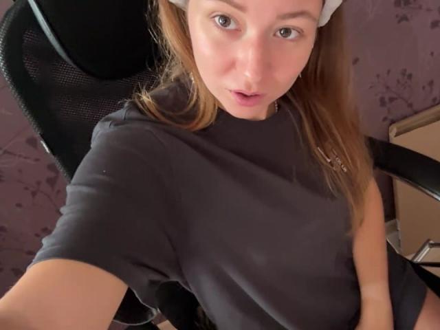 Ardelia Amesbury, com pele clara, olhos azuis e cabelo castanho preso em uma faixa branca, vestindo uma camisa preta, sentada em uma cadeira preta, olhando para a câmera com a boca ligeiramente aberta..