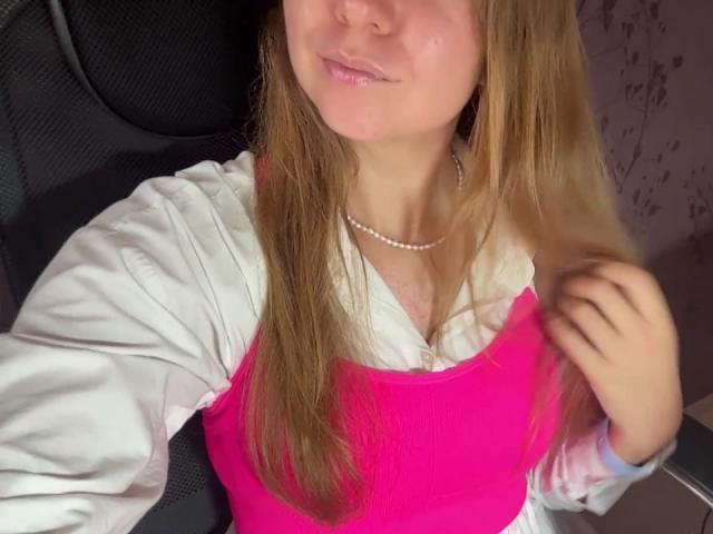 Ardelia Amesbury tiene el pelo largo y rubio, una camiseta sin mangas rosa y una camisa blanca con mangas abullonadas. Lleva un collar de perlas y una ligera sonrisa. Se ven los senos..