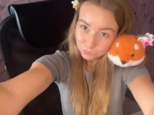 Ardelia Amesbury, con cabello rubio largo, vestida con una camisa gris, haciendo pucheros, tiene un peluche de un zorro naranja esponjoso con una flor rosa en el hombro, en un selfie..