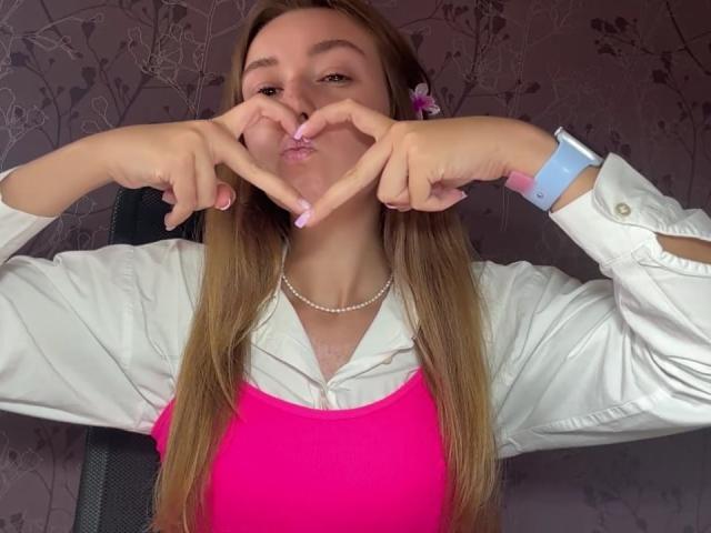 Ardelia Amesbury, com cabelos castanhos longos, blusa rosa, camisa branca, colar de pérolas, fazendo um formato de coração com os dedos, usando um smartwatch azul, presilha de cabelo rosa..