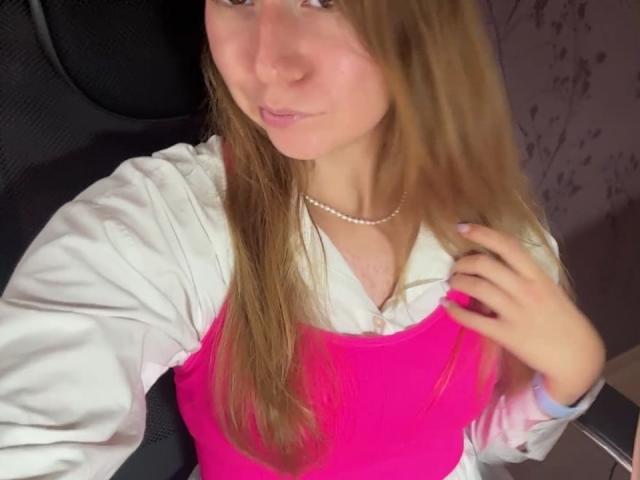 Ardelia Amesbury mit langen braunen Haaren, einem weißen Hemd und einem pinken Oberteil, das ihre kleinen Brüste zeigt, trägt eine Perlenkette, sitzt auf einem schwarzen Stuhl und lächelt in die Kamera..