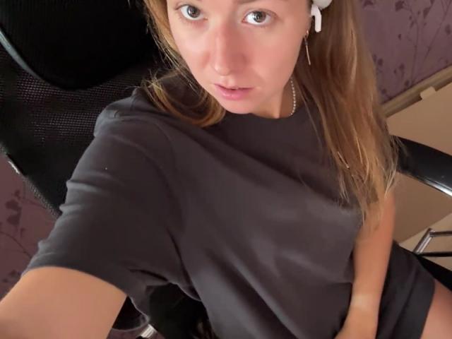Ardelia Amesbury, con cabello castaño claro, vestida con una camisa negra, sentada en una silla, mirando hacia arriba con un ligero ceño fruncido, sosteniendo su camisa cubriendo sus pequeños senos..
