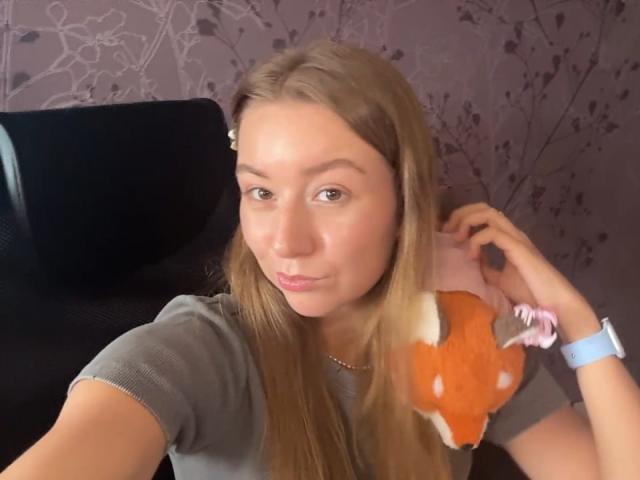 Ardelia Amesbury, com cabelos loiros longos, vestindo uma camisa cinza, segurando um brinquedo de raposa de pelúcia rosa e laranja, sentada em uma cadeira preta, olhando para a câmera, com um sorriso leve..