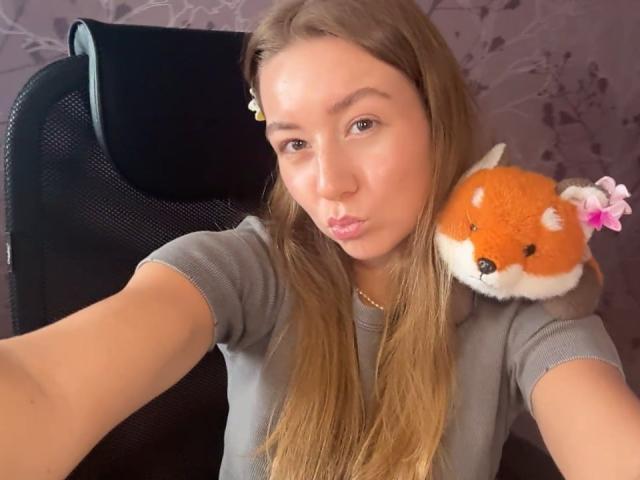 Ardelia Amesbury, con labios carnosos, sosteniendo un peluche de un zorro naranja esponjoso con flores rosas, vestida con una camisa gris, sentada en una silla de malla negra..
