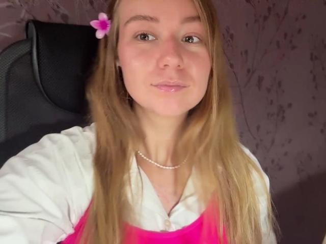 Ardelia Amesbury tiene el cabello largo y rubio, una flor rosa en el pelo, y lleva una camisa blanca con un top rosa, sonriendo suavemente con un collar de perlas..