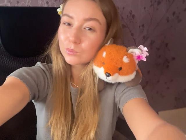 Ardelia Amesbury, con cabello rubio largo, una camisa gris y un juguete de zorro naranja esponjoso con una flor rosa en el hombro, frunciendo sus labios ligeramente rosados en un selfie..