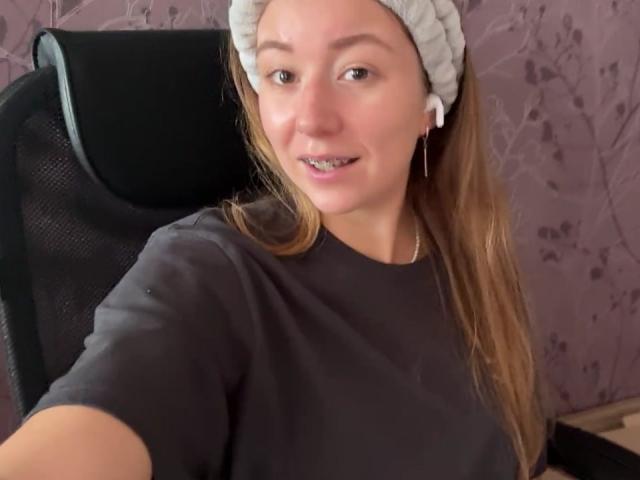 Ardelia Amesbury, con un gorro blanco de punto, una camisa negra y auriculares, sonriendo con su cabello castaño suelto, sentada en una silla de oficina negra de malla..