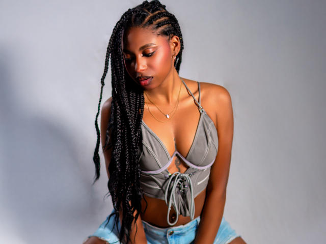 QueenPerlaa mit langen schwarzen Zöpfen, einem grauen Top, das ihre Brüste zeigt, und Jeansshorts, die nach unten blickt und dabei ein leichtes Lächeln hat, steht vor einem grauen Hintergrund..