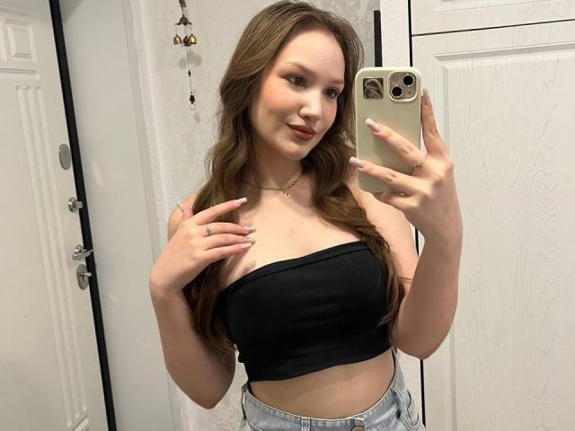 ShaundaOleksy, con lunghi capelli castani, indossa un top nero senza spalline, che mostra il seno, tiene un telefono, sorride, in un bagno con porte bianche..