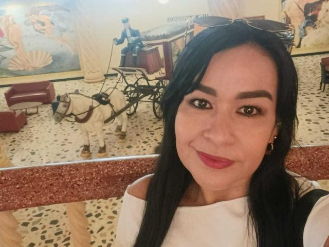 TamaraFoxy, con lunghi capelli neri, indossa una maglietta bianca con una spalla scoperta, sorride alla telecamera, ha piccoli occhi marroni, rossetto rosso e orecchini dorati. È in piedi di fronte a una carrozza trainata da cavalli..
