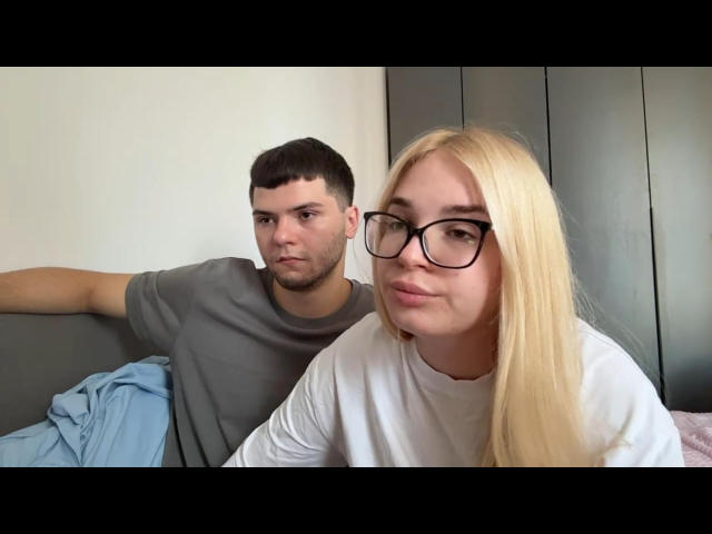 Eine blonde Frau namens Lessie Kirtner, mit Brille, weißem Hemd und kleinen Brüsten, sitzt neben einem dunkelhaarigen Mann in einem grauen Hemd auf einem Bett mit einer blauen Decke..