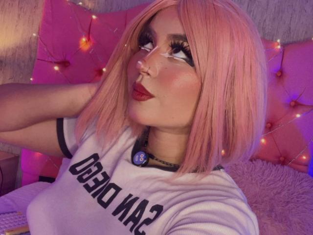 HakiraDiLucas mit einem pinken Bob, starkem Eyeliner und rotem Lippenstift, trägt ein weißes Hemd mit schwarzem Besatz, hat geschwollene Lippen und eine Halskette mit einem blauen Knopf. Brüste sind sichtbar..