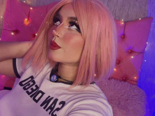 HakiraDiLucas, avec une coupe de cheveux courte rose, un trait d'eye-liner prononcé et du rouge à lèvres rouge, faisant la moue, portant une chemise blanche avec des détails noirs, et une épingle bleue. Guirlandes lumineuses violettes et roses..