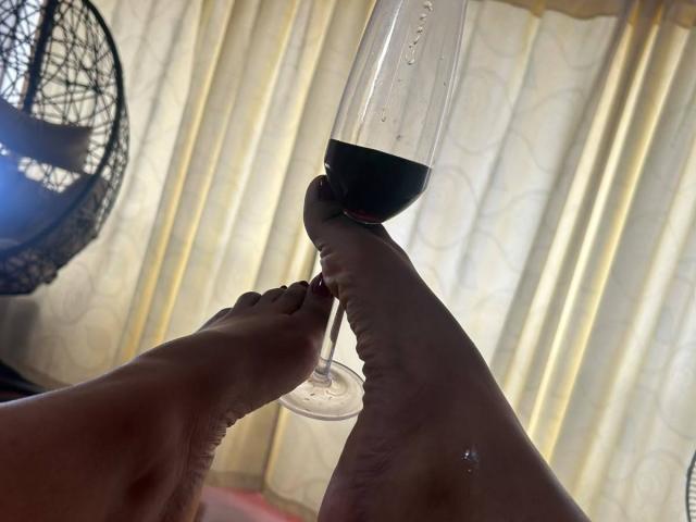 De voeten van NatalyBlonder houden een glas rode wijn vast, haar tenen raken de steel, met een lichte reflectie op de wijn. De zool van haar voet heeft een kleine waterdruppel..