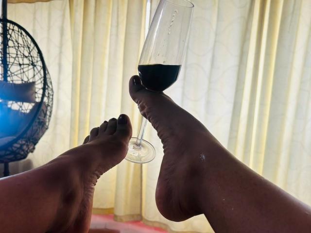 De voeten van NatalyBlonder, met donkere zolen en zwarte teennagels, raken een champagneglas met donkerrode wijn aan, in een ontspannen, intieme setting, met een rieten stoel op de achtergrond..
